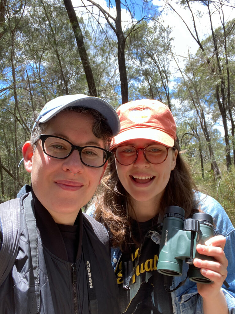 Two people facing the camera out in the bush, wearing caps, glasses and binoculars.