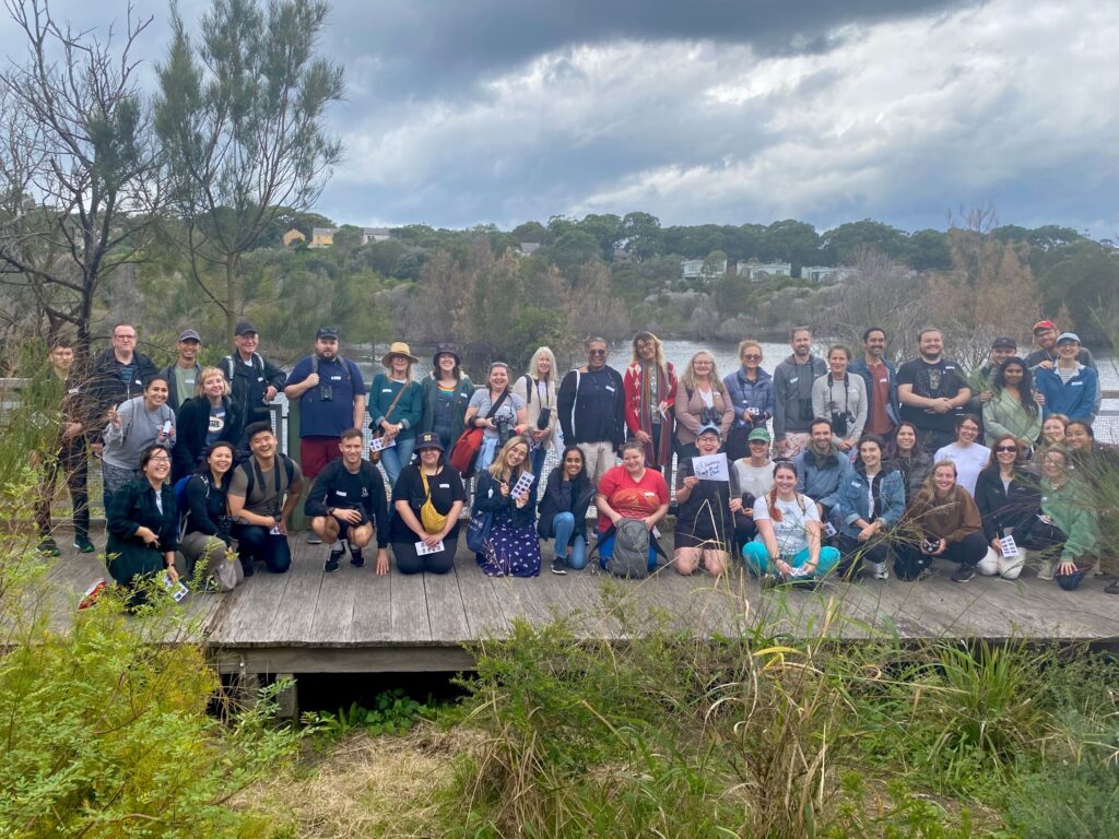 A large group of birdwatchers outdoors and posing for the camera. They are standing and crouching on a boardwalk in front of water.