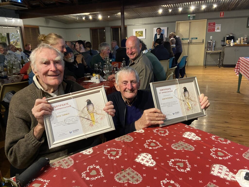 In a crowded hall, two men are seated at a table with a colourful red tablecloth, holding framed certificates with Regent Honeyeaters and smiling at the camera.