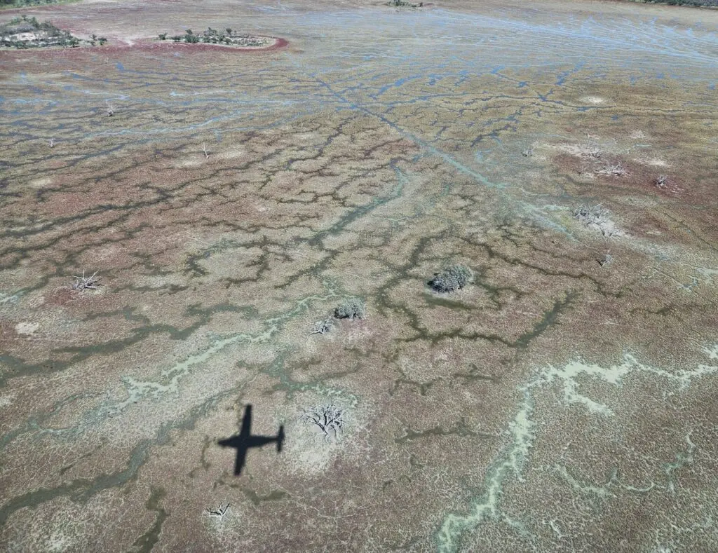 An aerial view of a drying pink, green and blue lake basin. The shadow of a plane is visible to the lower left of the frame. 